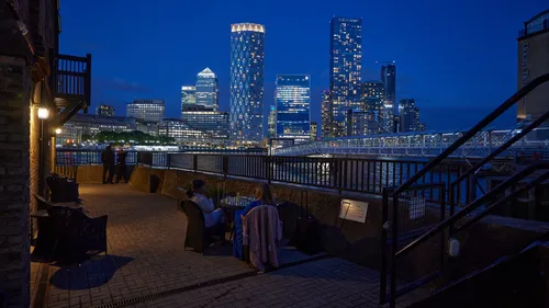 Canary Wharf Dusk Thames restaurant 2024 This urban architecture photograph captures an evening scene at a Thames-side restaurant in London during the summer season, with the main subject being the Canary Wharf riverside dining area. The image shows people seated at outdoor tables overlooking the River Thames, while others stand near the railing enjoying the view. Illuminated modern skyscrapers, including One Canada Square and the distinctive cylindrical Newfoundland Tower, dominate the skyline of Canary Wharf in the background. The lights from the buildings are reflected on the river, highlighting the vibrant atmosphere of London’s financial district at dusk. The overall composition emphasizes the blend of contemporary architecture and riverside urban life along the Thames.
